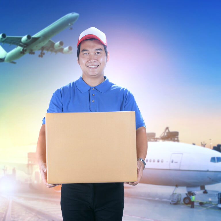 delivery man holding card box toothy smiling face against shipping port and cargo plane flying background